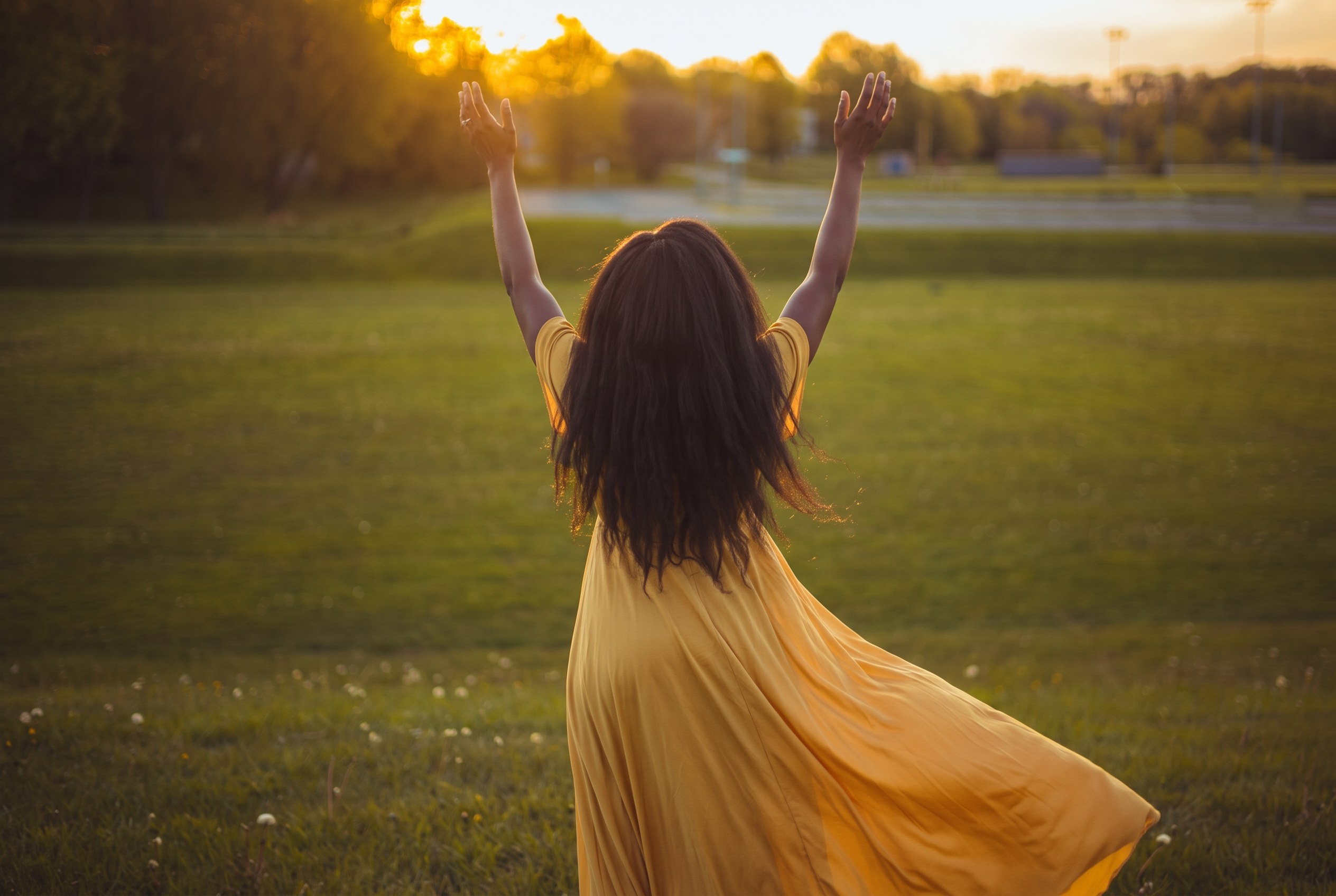 A woman in a flowing yellow dress lifting her hands in worship in a meadow at sunset